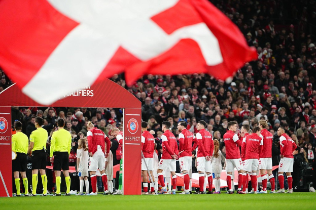 Eine Gruppe von Menschen auf einem Fußballfeld mit einer roten und weißen Flagge im Vordergrund, einem Bogen mit der Aufschrift "Bayern München vs Bayern München Wett-Tipps & Vorschau" im Hintergrund und einer großen Menge im Stadion.