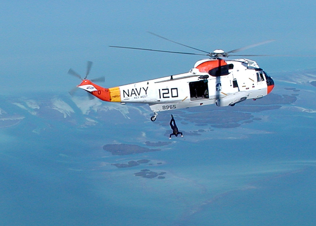 Hubschrauber fliegt über Wasser mit einer Person darunter, als Rettungshubschrauber etikettiert, vor einem Himmelhintergrund.