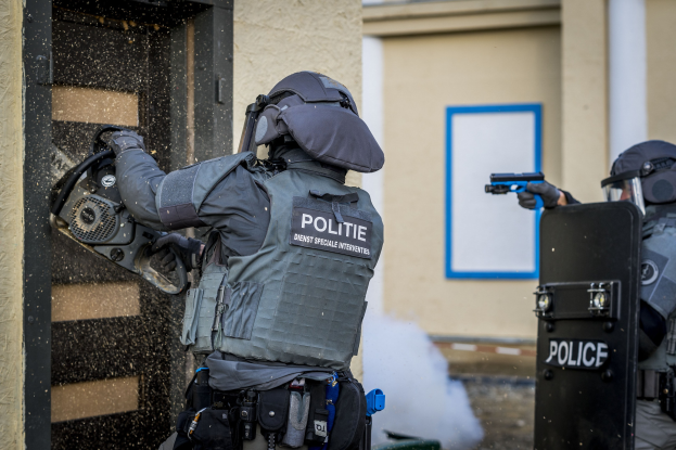 Zwei Polizisten in Einsatzausrüstung stehen vor einem Gebäude, einer hält eine Waffe und der andere ein Objekt, mit einer Tür links und einem vernagelten Fenster im Hintergrund.