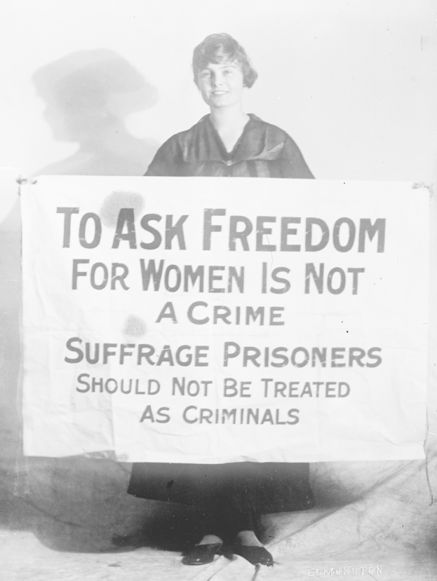 Schwarz-weiß-Foto einer Frau, die ein Banner hält, auf dem steht: "Für die Freiheit der Frauen zu kämpfen ist kein Verbrechen, Suffragetten sollten nicht wie Kriminelle behandelt werden." und dabei lächelt.