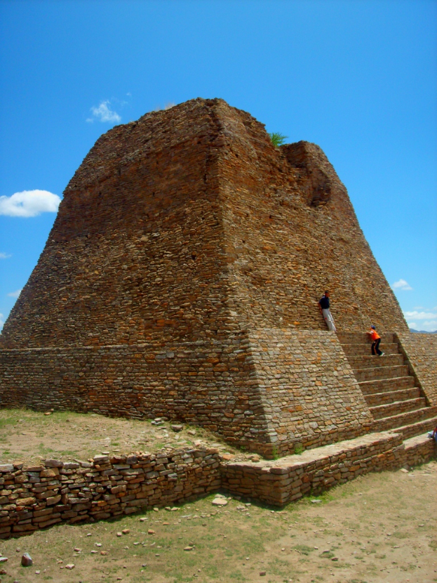 Eine archäologische Struktur mit Gras am Boden, ein Junge auf der Treppe rechts daneben, ein Mann auf der Struktur stehend und der Himmel darüber.