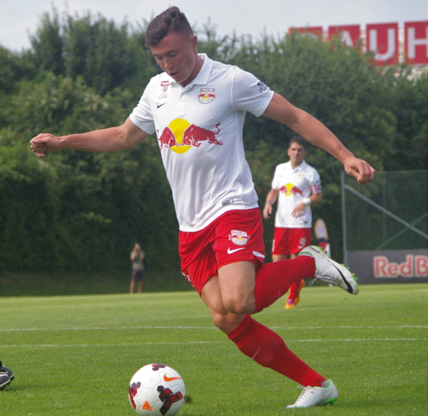 Ein Mann in einem Fussballtrikot schießt einen Ball auf einem grünen Feld mit Bäumen und einem klaren blauen Himmel im Hintergrund, mit einem Schild, auf dem "RB Leipzig" zu sehen ist.