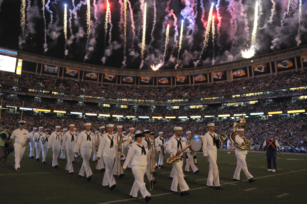 Gruppe von Menschen in weißen Uniformen marschiert auf einem Fußballfeld, einige halten Musikinstrumente, mit einer großen Menge im Stadion, Bannern, Bildschirmen links und Feuerwerk am Himmel.