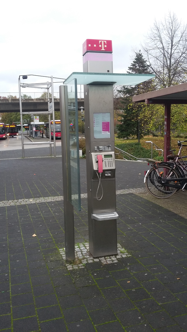 Eine Telefonzelle steht an der Straße neben einer Bushaltestelle, daneben parkt ein Fahrrad; im Hintergrund sind Bäume, Fahrzeuge auf der Straße, eine Brücke und ein klarer blauer Himmel zu sehen.