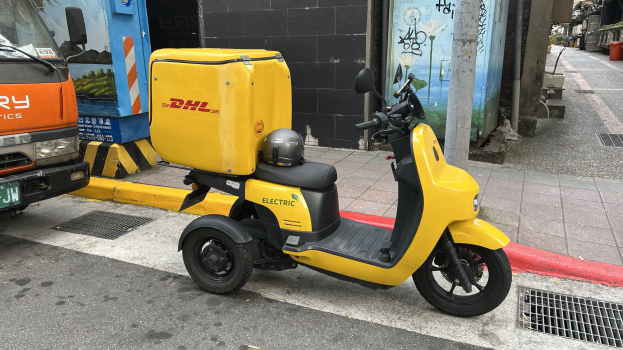 Ein gelbes Elektro-Scooter mit Helm darauf steht auf der Straße neben einem Lastwagen, mit Gebäuden und Menschen auf einem Fußweg im Hintergrund.