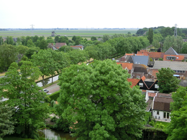 Eine Stadtrandszene mit Häusern und Bäumen, Autos auf einer Straße und Türmen mit einer Windmühle im Hintergrund.