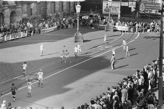 Schwarzes Bild eines Straßenfußballspiels mit Spielern und Zuschauern, Laternen, Gebäuden und Bannern im Hintergrund.