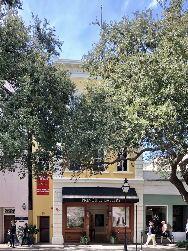 Außenansicht der Principle Gallery in Charleston, SC, mit Menschen auf dem Bürgersteig, Topfpflanzen, einer Straßenlampe, einem Schild, einer Fahne, einem Fahnenmast und einem bewölkten Himmel.