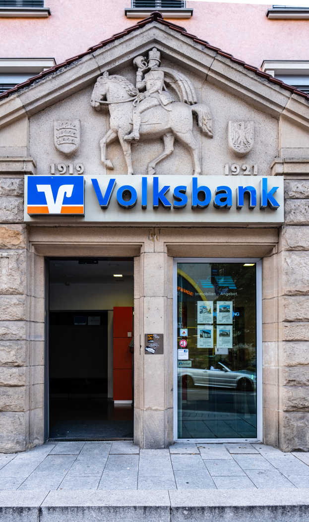 Ein als Volksbank identifiziertes Gebäude in Frankfurt, Deutschland, mit Fenstern, einer Tür mit Plakaten, einer Texttafel und einer Skulptur eines Menschen auf einem Pferd, mit einem Weg, der zum Gebäude führt.