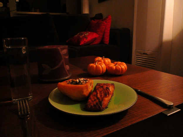 Ein Holztisch mit Kürbissen, ein Teller mit Essen, ein Glas, ein Becher und Besteck, vor einem Sofa mit Kissen, vor einer Wand mit einer Tür auf der rechten Seite.