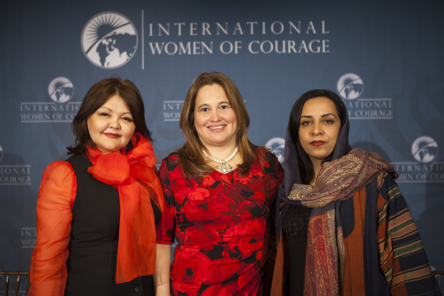 Drei Frauen, die fröhlich vor einem Banner mit der Aufschrift 'Internationale Frauen des Muts' stehen.