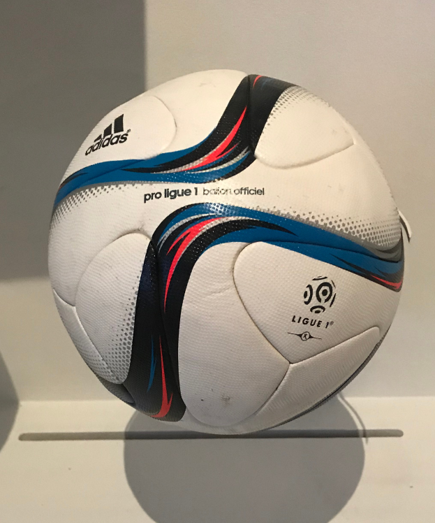 Ein weißer Fußball mit dem Adidas-Logo und 'Ligue 1' in fetter schwarzer Schrift auf einem weißen Tisch auf einem hellen weißen Hintergrund.