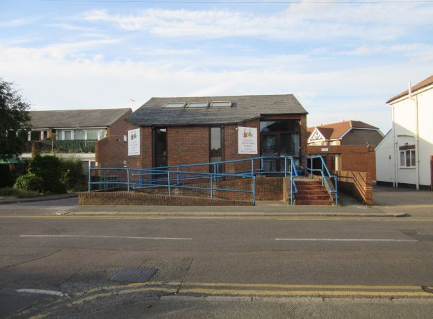 Ein Schulgebäude mit blauer Begrenzung, umgeben von Häusern, Treppen, Geländern, Texttafeln, Pflanzen, Bäumen und einem bewölkten Himmel.