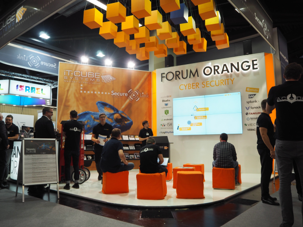 Eine Gruppe von Menschen an einem IT-Cube Systems Forum Orange Cyber Security Stand auf einer Messe, mit sitzenden und stehenden Teilnehmern, einem Bildschirm, Texttafeln, Regalen mit Gegenständen und Deckenbeleuchtung.