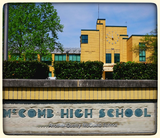Ein Plakat mit der Aufschrift "MCCOMB HIGH SCHOOL" an einer Wand, mit Pflanzen, Bäumen, einem Pfahl, einem Gebäude und einem bewölkten Himmel im Hintergrund.