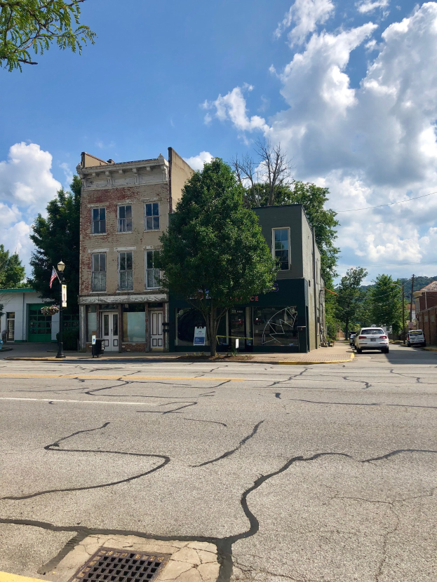 Leere Straße mit parkenden Autos, Gebäuden, Bäumen, Laternenmästen und einem bewölkten Himmel; eine beschädigte Straßenrück und ein feuerbeschädeltes Gebäude im Vordergrund.