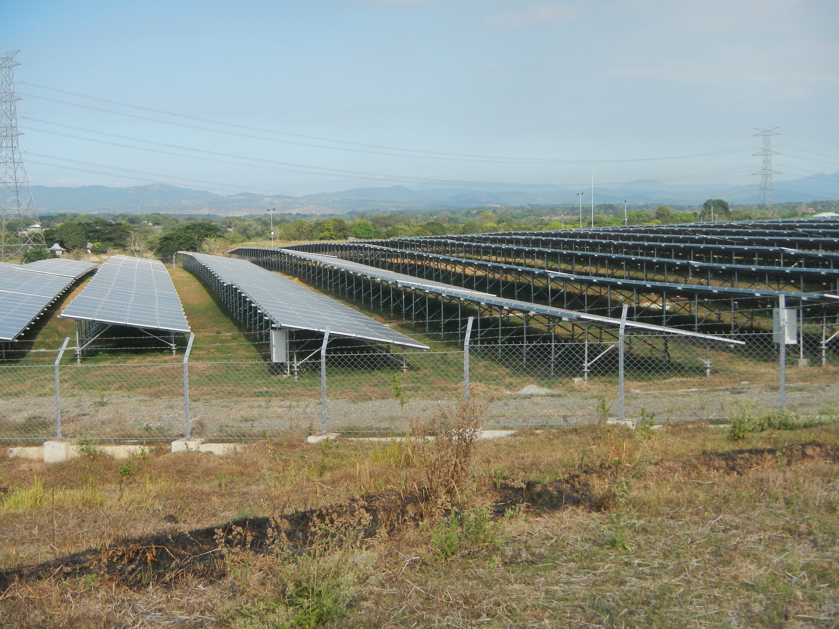 Luftaufnahme eines Solarpanelfelds, das von einem Zaun, Gras und Pflanzen umgeben ist, mit Bäumen, Übertragungstürmen, Stromleitungen und Hügeln unter einem klaren blauen Himmel.