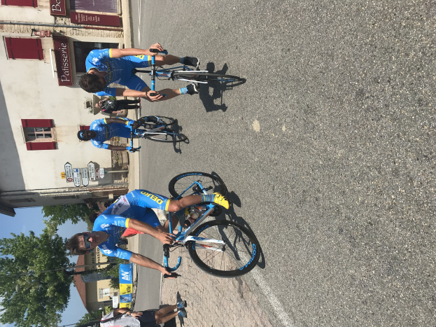 Eine Gruppe von Radfahrern fährt eine Straße entlang an einem Gebäude mit vernagelten Fenstern und Text vorbei, mit Bäumen und einem blauen Himmel im Hintergrund und Zuschauern auf der Straße darunter.