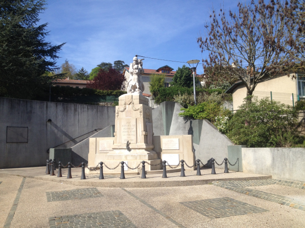 Statue eines Mannes auf einem Sockel in einem Park, umgeben von Bäumen, Pflanzen, einem Metallzaun, einem Straßenpfahl und Gebäuden unter einem bewölkten Himmel, angeblich ein Denkmal für die Pariser Anschläge.