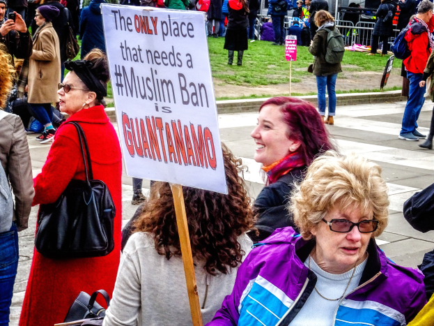 Eine Gruppe von Menschen steht auf dem Boden, einige halten Taschen, eine Frau hält ein Schild mit der Aufschrift "Der einzige Ort, der ein Muslimverbot benötigt, ist Guantanamo". Im Hintergrund sehen wir einen Metallzaun, Gras, ein Schild mit Text, einige Pfosten, ein Gebäude mit Fenstern und Bäume.