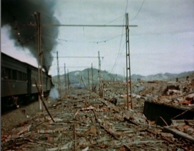 Ein Zug fährt durch ein beschädigtes Gebiet mit Rauch, der aus ihm aufsteigt, umgeben von Strommasten und -kabeln, mit Hügeln und einem klaren Himmel im Hintergrund und verstreuten Schrottteilen auf dem Boden.