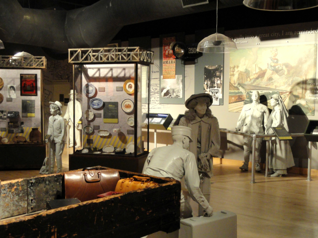 Ein Museumsraum mit verschiedenen Artefakten, darunter Puppe mit Hut, Koffer, Kiste und andere Gegenst├Ąnde auf dem Boden, sowie Informationsschilder an den W├Ąnden und Lampen an der Decke.