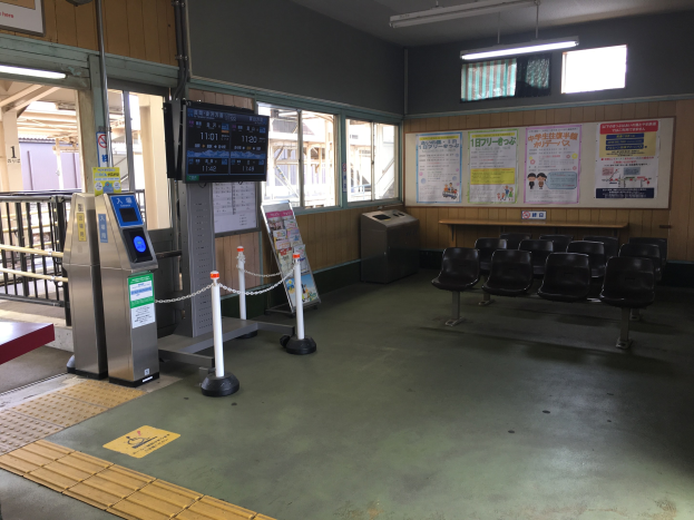 Ein belebter Bahnhofseingang mit Reihen von Stühlen, einer Service-Theke, Ticketautomaten, Kettenpfählen, Informationsanzeigen, Wandplakaten, Deckenbeleuchtung und Geländern.