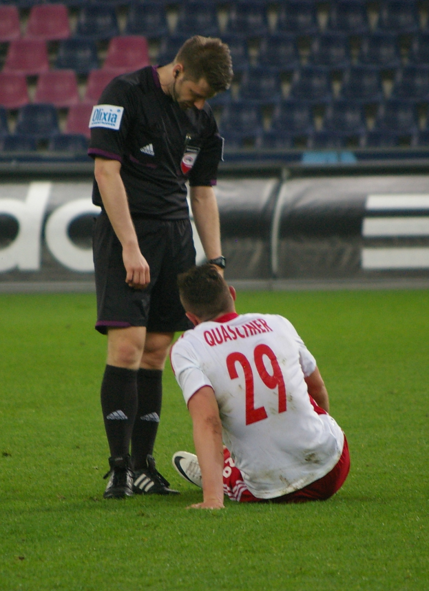 Ein Füßballspieler sitzt neben einem Schiedsrichter auf dem Boden, beide in Sportkleidung, mit Tafeln und Stühlen im Hintergrund eines Stadions.