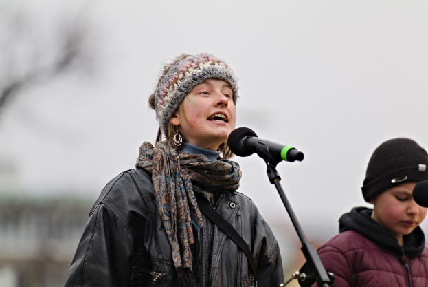Eine Frau in schwarzer Jacke, Schal und Mütze singt in ein Mikrofon auf der Bühne, neben einer Person in einer maronenfarbenen Jacke und schwarzer Mütze, während der unscharfe Hintergrund einen träumerischen Effekt erzeugt.