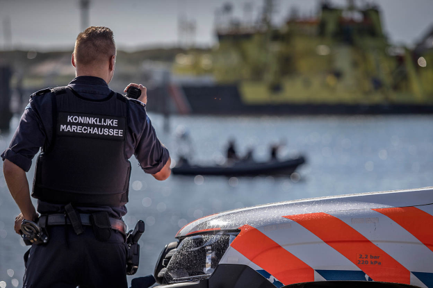 Ein Polizist in Uniform steht neben einem Polizeiwagen in der Nähe eines Gewässers und hält ein Funkgerät in der Hand, im Hintergrund ist ein Boot mit ein paar Menschen auf dem Wasser zu sehen und der Himmel darüber.