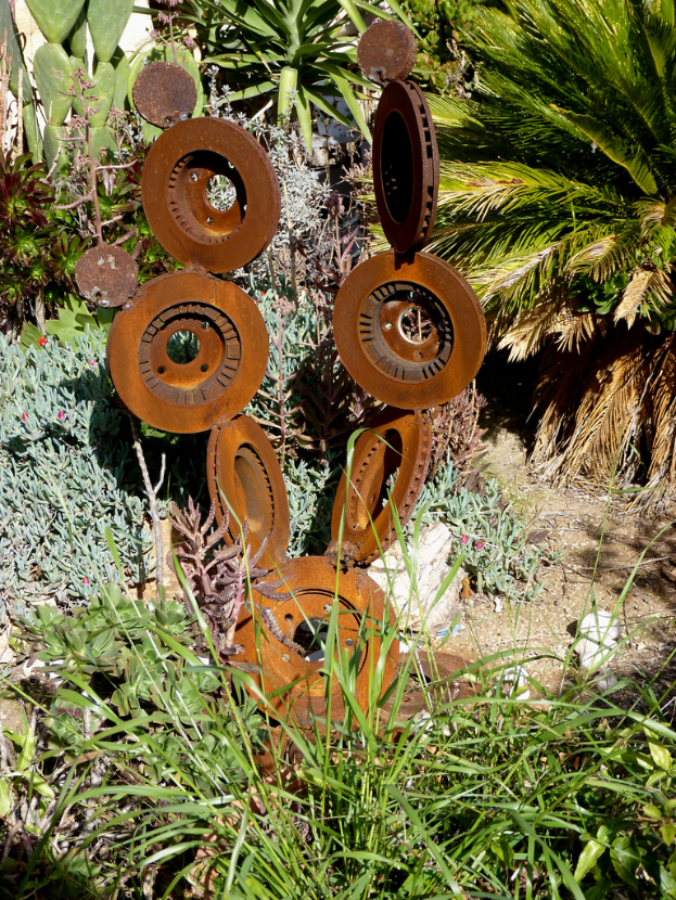 Eine Metallskulptur steht auf einem Stück Land mit spärlicher Vegetation, einschließlich einiger Pflanzen, Grasflecken und verstreuten Bäumen.