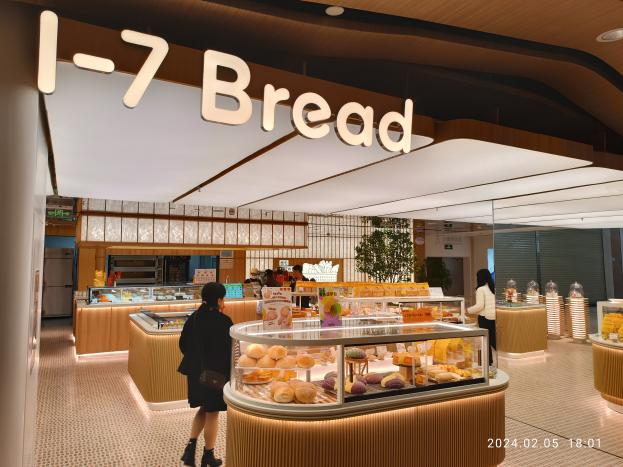 Eine Frau steht vor einer Bäckertheke, die mit 'I-7 Brot' beschriftet ist und zeigt verschiedene Lebensmittel, umgeben von ein paar Menschen und einer Pflanze im Hintergrund.