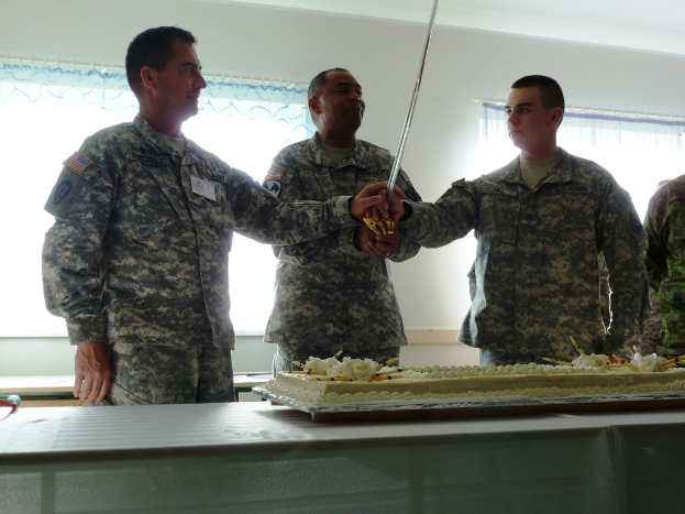 Ein großer Kuchen auf einem Tisch mit mehreren Soldaten davor, drei halten ein großes Messer und eine Wand mit zwei Fenstern im Hintergrund.