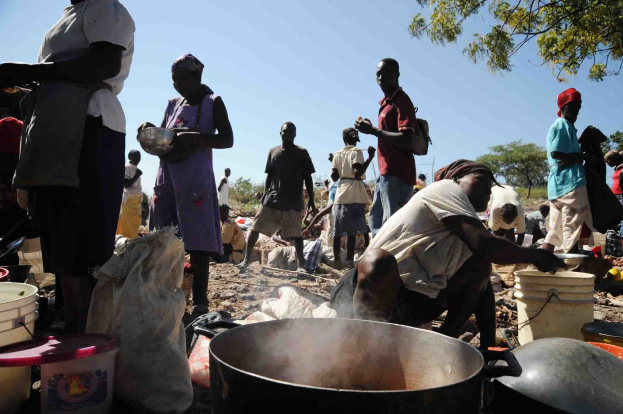 Eine Gruppe von Menschen, die in einer l├Ąndlichen Gegend im Freien Essen zubereiten, mit einem gro├čen Topf, Eimern und anderen Gegenst├Ąnden auf dem Boden, unter einem klaren blauen Himmel.