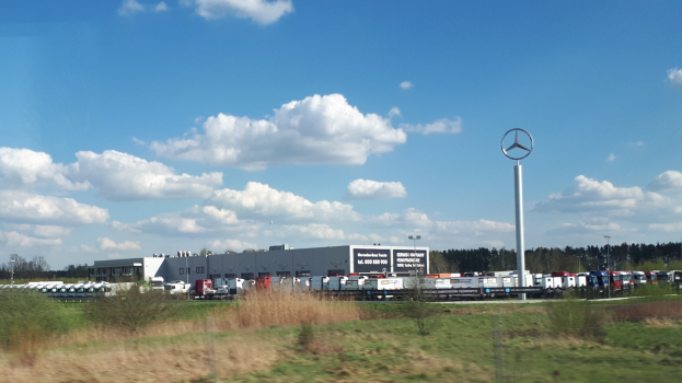 Ein Mercedes-Benz-Werk in Deutschland mit Fahrzeugen auf der Straße, Grünflächen, Gebäuden und einem Logo-Mast unter einem bewölkten Himmel.
