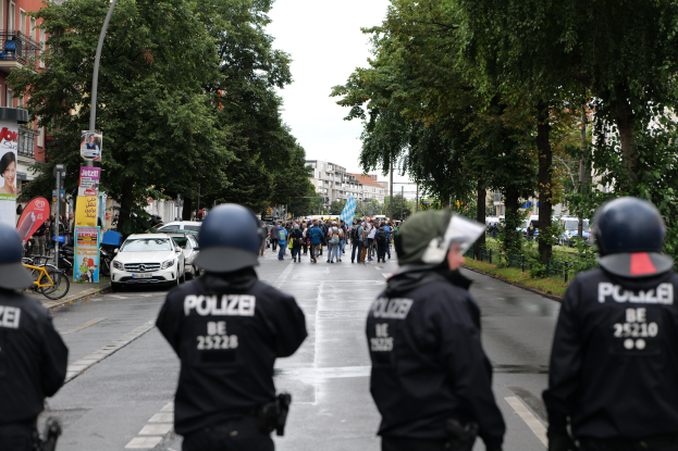 Eine Gruppe von Polizisten in schwarzen Uniformen und Helmen steht an der Seite einer Straße, mit Menschen, Fahrzeugen, Fahrrädern, Pfählen, Brettern, Bäumen, Gebäuden und einer Fahne im Hintergrund.