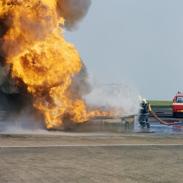 Feuerwehrfahrzeug in Flammen auf der Seite einer Straße, mit zwei Helmen tragenden Personen, die Rohre halten, und einem Fahrzeug im Hintergrund.