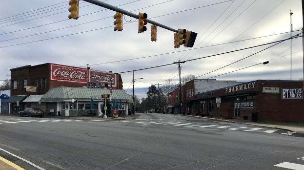 Stadtstraßenkreuzung mit Ampel, Fahrzeugen, Gebäuden mit Schildern, Strommasten und -leitungen, Bäumen und einem bewölkten Himmel.
