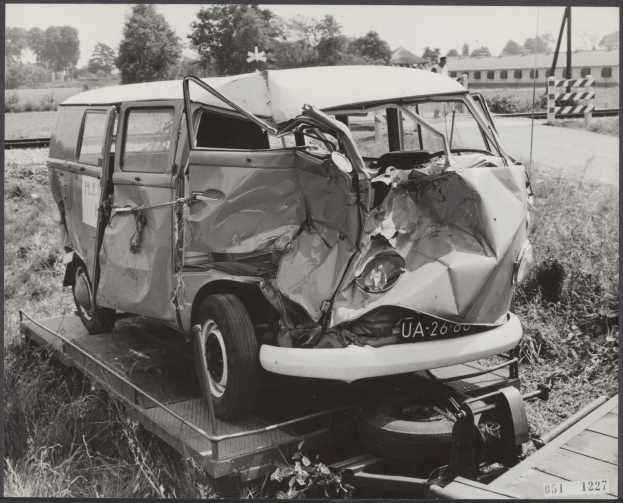Schwarz-weißes Foto eines schwer beschädigten Autos mit eingedellter Front und zersplitterten Fenstern auf einem Tieflader, umgeben von Gras und Pflanzen mit Bäumen und einem Gebäude im Hintergrund.