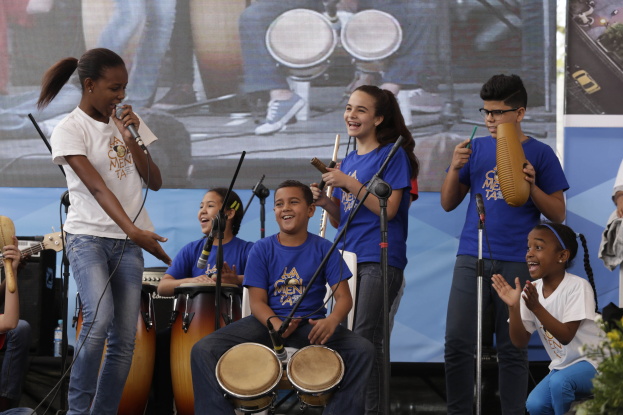 Kinder mit Musikinstrumenten auf einer Bühne mit einer Frau mit Mikrofon, umgeben von Mikrofonstativen, einem Banner im Hintergrund und Pflanzen auf der rechten Seite.