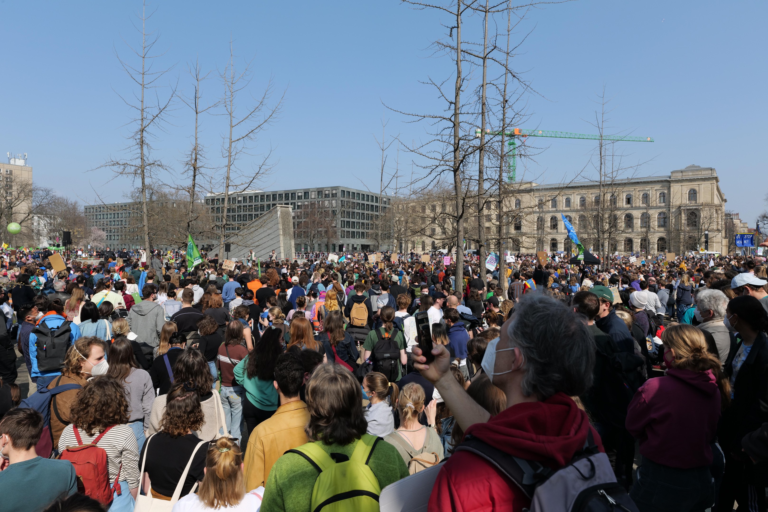 Große Menschenmenge mit Schildern und Taschen vor einem Gebäude mit Bäumen und einem klaren Himmel, was auf eine Klimaprotestveranstaltung hinweist.