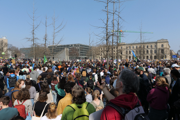 Große Menschenmenge mit Schildern und Taschen vor einem Gebäude mit Bäumen und einem klaren Himmel, was auf eine Klimaprotestveranstaltung hinweist.