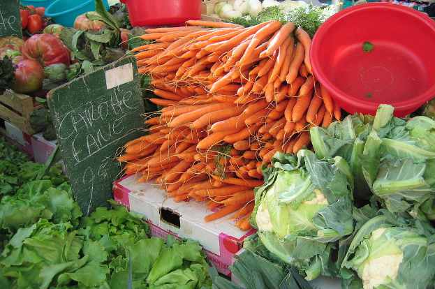 Eine MarktSzene mit verschiedenen Gemüsen wie Karotten, Blumenkohl, Blattgemüse, Zwiebeln und anderen, die in Körben und Kartons displayed sind, mit Text auf Holztafeln im Hintergrund.