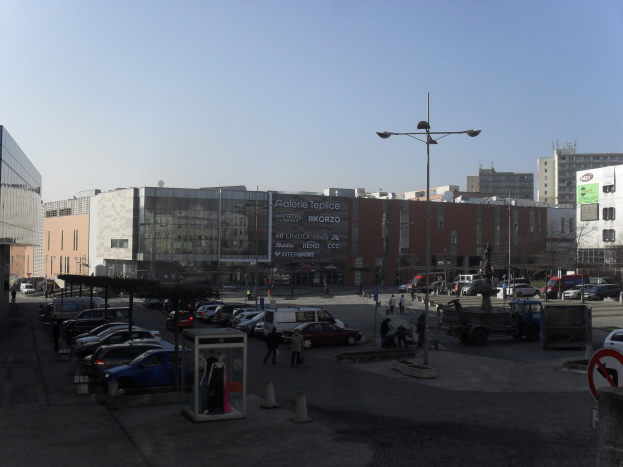 Eine belebte Stadtstraße mit parkenden Autos auf einem Parkplatz neben hohen Gebäuden, beleuchtet von Straßenlaternen, mit einigen Fußgängern und sichtbarem Himmel im Hintergrund.