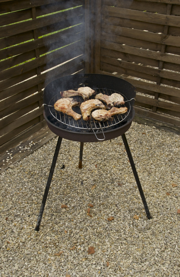 Grill mit Fleisch darauf auf einem Kiesgelände, umgeben von einem Holzzaun und Bäumen, mit Rauch, der aus dem Grill aufsteigt.