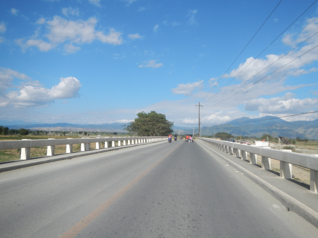Eine Autobahn mit Motorradfahrern, umgeben von Geländern, Strommästen mit Kabeln, Bäumen, Hügeln und einem bewölkten Himmel.