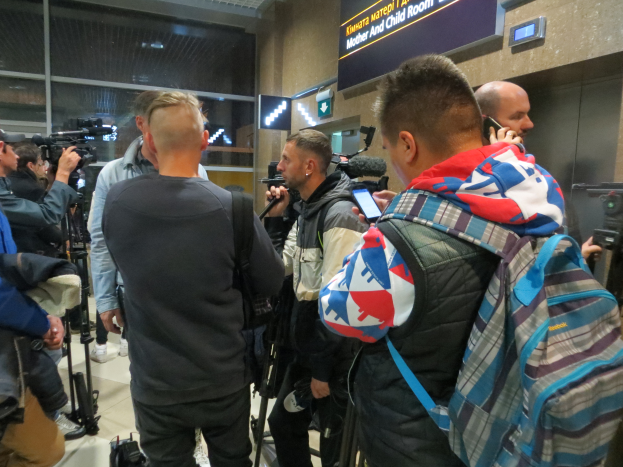 Eine Gruppe von Menschen, einige mit Kameras und Handys, andere mit Taschen, steht in einem Flughafen-Pressebereich mit einem Textschild und Lampen dahinter.
