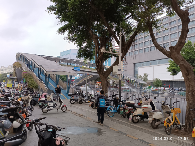 Eine belebte Stadtstraße mit parkenden E-Scootern, Fußgängern auf dem Gehweg, Bäumen, Gebäuden mit Fenstern und einem bewölkten Himmel, mit einem Wasserzeichen.