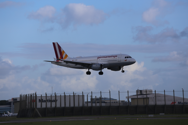 Ein Airbus A320-200 der Germanwings beim Start vom Frankfurter Flughafen, mit Gras, einem Zaun, Gebäuden, Bäumen und einem bewölkten Himmel im Hintergrund.