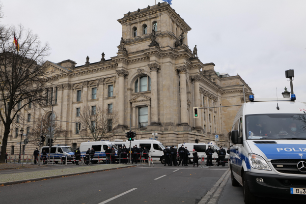 Eine Gruppe von Polizeibeamten steht vor dem Reichstaggebäude in Berlin, Deutschland, mit Fahrzeugen, einem Zaun, Verkehrsampeln, Laternenpfählen, Bäumen und Fahnen im Hintergrund, bei klarem Himmel.